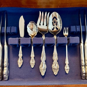 May include: A set of ornate silver-colored flatware, including knives, forks, and spoons, arranged in a blue velvet-lined wooden case. The silverware features detailed floral patterns on the handles. The set includes a serving spoon and a butter knife.