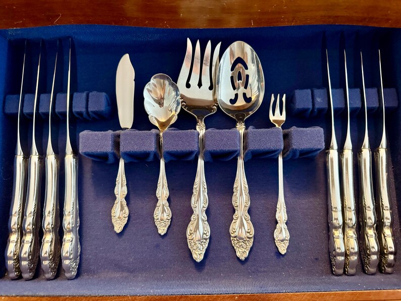 May include: A set of ornate silver-colored flatware, including knives, forks, and spoons, arranged in a blue velvet-lined wooden case. The silverware features detailed floral patterns on the handles. The set includes a serving spoon and a butter knife.