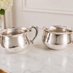 Puede incluir: Dos azucareros de plata pulida con bordes festoneados y asas ornamentadas y curvadas. Cada cuenco presenta un cuerpo redondeado y abombado con un acabado met&aacute;lico reflectante, mostrando detalles intrincados en las asas y una boca ancha.