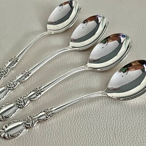 May include: Four silver spoons with floral detailing on the handles. The spoons have a reflective, polished surface, and are arranged diagonally across a light-colored surface. The spoons are designed for serving.