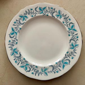 Op de afbeelding: Een wit keramieken bord met een geschulpte rand en een gouden metalen rand. De rand is voorzien van een herhalend patroon van blauwgroene en grijze bladeren, krullende ranken en kleine blauwgroene stippen in een cirkelvormig ontwerp.