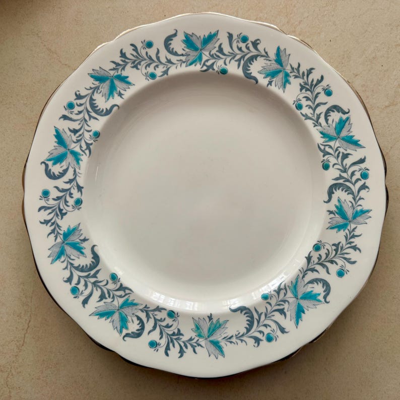 Op de afbeelding: Een wit keramieken bord met een geschulpte rand en een gouden metalen rand. De rand is voorzien van een herhalend patroon van blauwgroene en grijze bladeren, krullende ranken en kleine blauwgroene stippen in een cirkelvormig ontwerp.