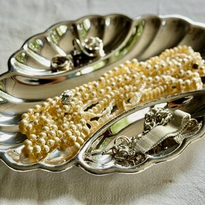 Pode incluir: Um prato de metal prateado em forma de concha com borda recortada e interior dividido. O prato cont&eacute;m um longo colar de p&eacute;rolas artificiais na cor creme e uma pulseira de corrente de metal prateado com um design de malha texturizada.