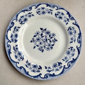 May include: A round ceramic plate with a cream-colored base featuring a dark blue floral pattern. The center displays a cluster of flowers, while the rim is decorated with a continuous scrolling border of blue flowers and intricate leaf designs.
