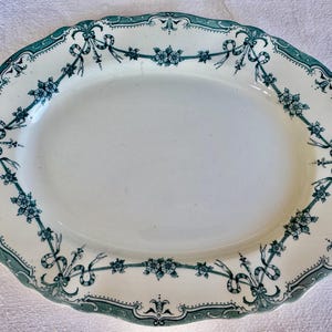 May include: An oval-shaped serving platter with a cream-coloured base and a scalloped edge. The rim features a dark green transferware pattern consisting of floral clusters connected by flowing ribbons and bows, creating a decorative border.