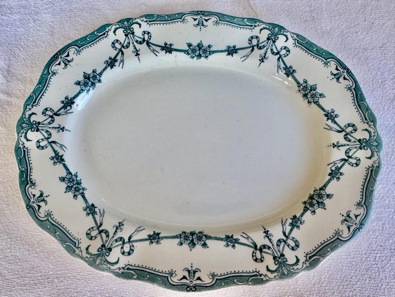 May include: An oval-shaped serving platter with a cream-coloured base and a scalloped edge. The rim features a dark green transferware pattern consisting of floral clusters connected by flowing ribbons and bows, creating a decorative border.