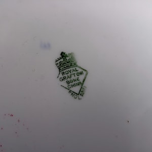 May include: A white ceramic surface featuring a green stamp with a crown icon above a diamond shape. The text inside the diamond reads ROYAL GRAFTON BONE CHINA ENGLAND. There are small red and blue ink marks visible near the stamp on the white background.
