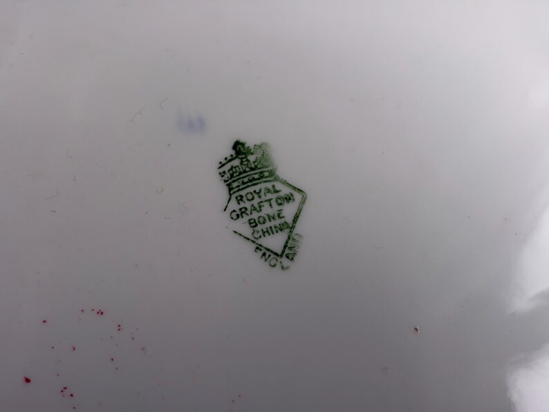 May include: A white ceramic surface featuring a green stamp with a crown icon above a diamond shape. The text inside the diamond reads ROYAL GRAFTON BONE CHINA ENGLAND. There are small red and blue ink marks visible near the stamp on the white background.