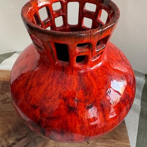 Puede incluir: Un jarr&oacute;n de cer&aacute;mica rojo vibrante con un dise&ntilde;o calado &uacute;nico en la parte superior. El jarr&oacute;n tiene un cuerpo bulboso y un acabado brillante, mostrando una mezcla de tonos rojos. La parte superior presenta una serie de cortes rectangulares.