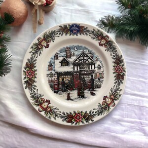 May include: A round ceramic plate featuring a central illustration of a snow-covered toy shop at night. The rim is decorated with a repeating pattern of pine cones, red flowers, and lanterns. A Royal Stafford sticker is at the top.
