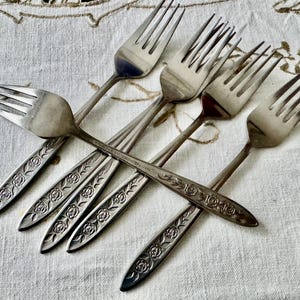 May include: A collection of six silver forks with a floral rose design on the handles. The forks are arranged in a crisscross pattern on a white tablecloth with a subtle pattern.