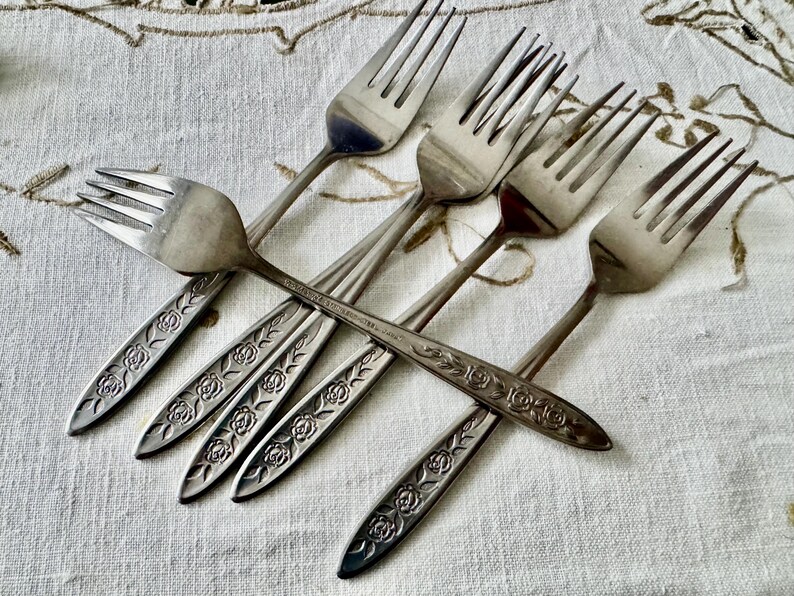 May include: A collection of six silver forks with a floral rose design on the handles. The forks are arranged in a crisscross pattern on a white tablecloth with a subtle pattern.