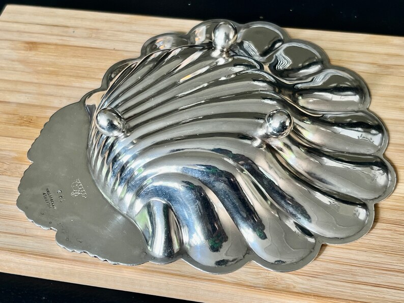 May include: A silver-plated serving dish shaped like a seashell with three feet. The dish has a scalloped edge and a smooth, shiny surface. The bottom of the dish is marked with the text "CRYSTAL  MADE IN  HONG KONG".