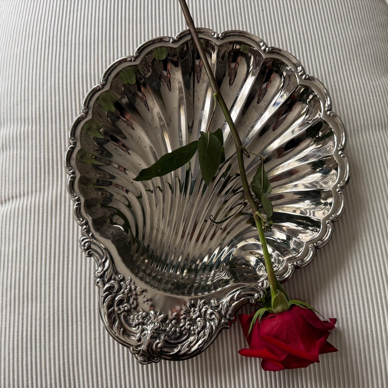 K&ouml;nnte beinhalten: Eine polierte, silberfarbene Metallschale in Form einer Jakobsmuschel mit gewelltem Rand und floralen Verzierungen an der Basis. Eine einzelne rote Rose mit gr&uuml;nen Bl&auml;ttern und einem langen Stiel liegt diagonal auf der Schale.