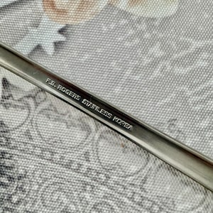 Może przedstawiać: Srebrzysty uchwyt sztućca ze stali nierdzewnej z wygrawerowanym napisem F.B. ROGERS STAINLESS KOREA. Metalowa powierzchnia ma gładkie, odblaskowe wykończenie z lekko wklęsłym środkowym rowkiem biegnącym wzdłuż całej długości elementu.