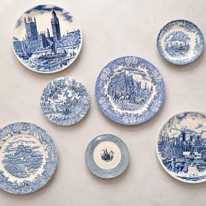 May include: A collection of six round ceramic plates featuring blue and white transferware patterns. The designs depict various architectural landmarks, rural landscapes, and intricate floral borders on a white background with blue ink.