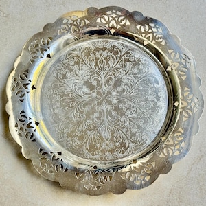 May include: A round silver-toned metal serving tray featuring a wide, intricate openwork filigree border with a scalloped edge. The center of the tray is smooth and decorated with a detailed, symmetrical floral and leaf engraving.