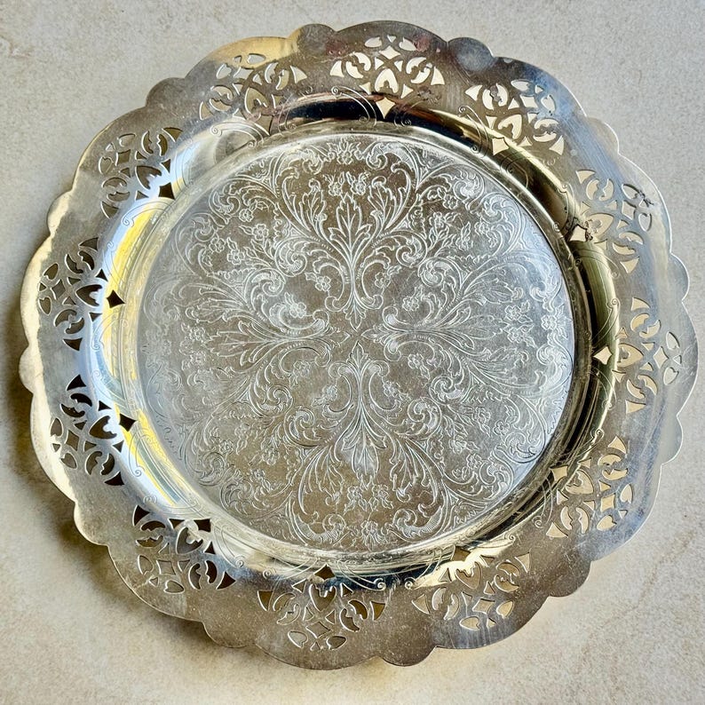 May include: A round silver-toned metal serving tray featuring a wide, intricate openwork filigree border with a scalloped edge. The center of the tray is smooth and decorated with a detailed, symmetrical floral and leaf engraving.