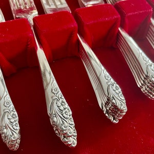 May include: Close-up of a set of silver silverware, including spoons and forks, with a floral design on the handles. The silverware is arranged on a red velvet surface, creating a striking contrast. The spoons and forks are neatly organized.