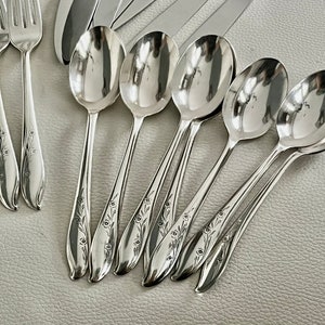 May include: A collection of polished silver flatware, including forks, spoons, and knives. The spoons are arranged in a row, with a floral design on the handles. The silverware is displayed on a light-colored surface.
