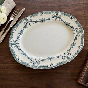 May include: An oval ceramic serving platter featuring a cream-coloured centre and a decorative border in teal and blue. The rim is adorned with a repeating pattern of floral garlands and ribbons. The platter has a scalloped edge and a smooth finish.
