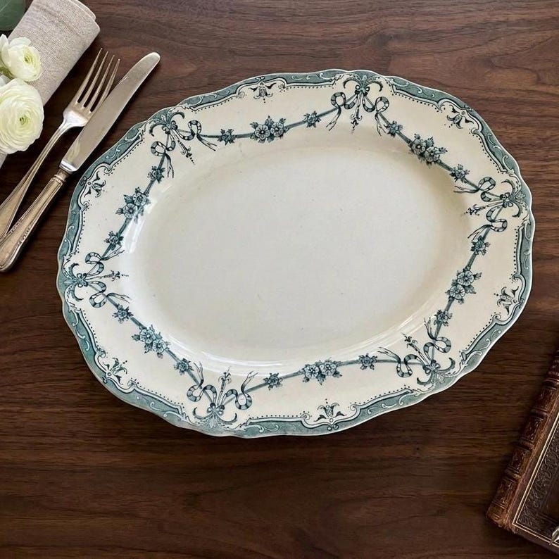 May include: An oval ceramic serving platter featuring a cream-coloured centre and a decorative border in teal and blue. The rim is adorned with a repeating pattern of floral garlands and ribbons. The platter has a scalloped edge and a smooth finish.