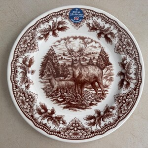 Op de afbeelding: Een wit keramieken bord met een bruine transferprint van een hert en een hinde in een bosrijke omgeving. De rand is versierd met een herhalend patroon van eikenbladeren en eikels. Een blauw-witte Royal Stafford sticker is op het bord aanwezig.