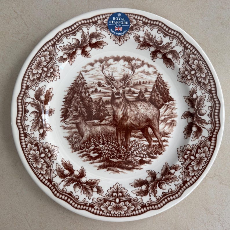 Op de afbeelding: Een wit keramieken bord met een bruine transferprint van een hert en een hinde in een bosrijke omgeving. De rand is versierd met een herhalend patroon van eikenbladeren en eikels. Een blauw-witte Royal Stafford sticker is op het bord aanwezig.