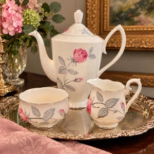 May include: A white porcelain tea set featuring a tall teapot, a creamer, and a sugar bowl, all decorated with pink rose blossoms and grey leaves. The pieces have silver-toned rims and are arranged on a round, ornate silver-toned metal tray.