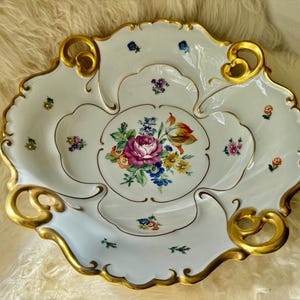 Puede incluir: Fuente de porcelana blanca con dise&ntilde;o floral y ribete dorado. La fuente presenta un ramo central de flores rosas y amarillas, rodeado de peque&ntilde;os detalles florales. Los bordes tienen un dise&ntilde;o festoneado y asas doradas.