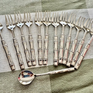 May include: A collection of silver-colored forks and a spoon with bamboo-style handles. The forks are arranged in two rows, with the spoon placed below. The silverware is set on a green and white checkered tablecloth.
