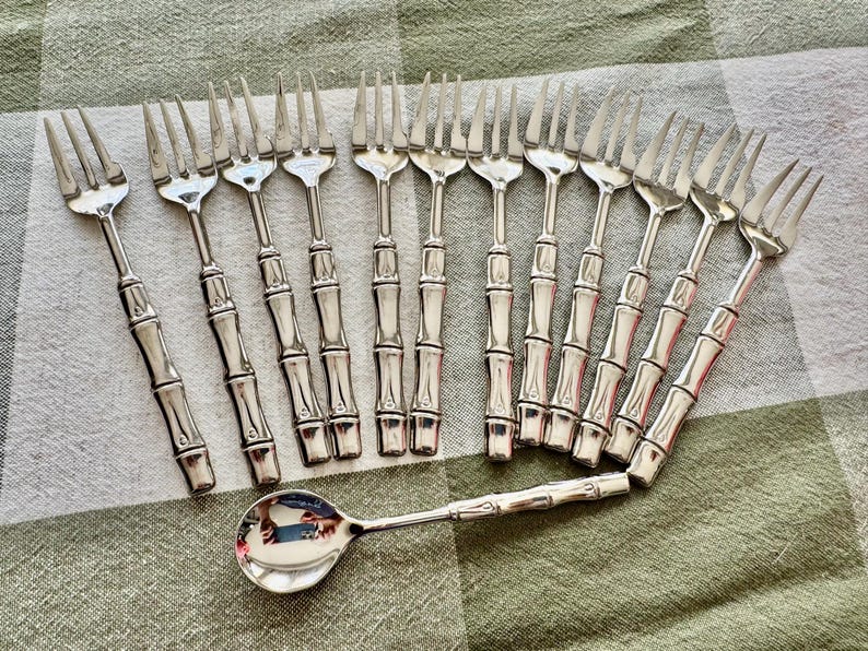 May include: A collection of silver-colored forks and a spoon with bamboo-style handles. The forks are arranged in two rows, with the spoon placed below. The silverware is set on a green and white checkered tablecloth.