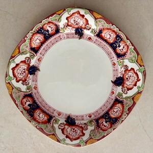 May include: A round ceramic plate featuring a central white basin surrounded by a decorative border. The rim displays a repeating pattern of orange and white floral motifs, dark blue accents, and light green scrollwork on a cream background.