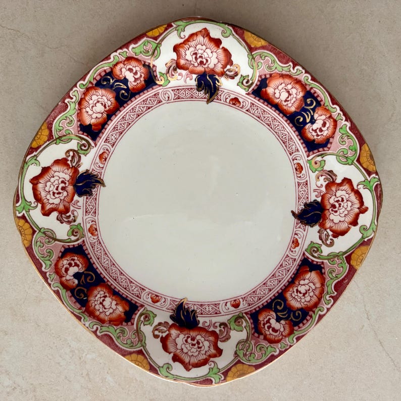 May include: A round ceramic plate featuring a central white basin surrounded by a decorative border. The rim displays a repeating pattern of orange and white floral motifs, dark blue accents, and light green scrollwork on a cream background.