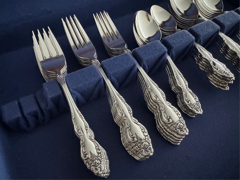 May include: Close-up of a set of ornate silverware, including forks and spoons, arranged in a blue case. The silverware has a detailed floral pattern on the handles and a shiny, reflective surface. The forks and spoons are neatly organized.