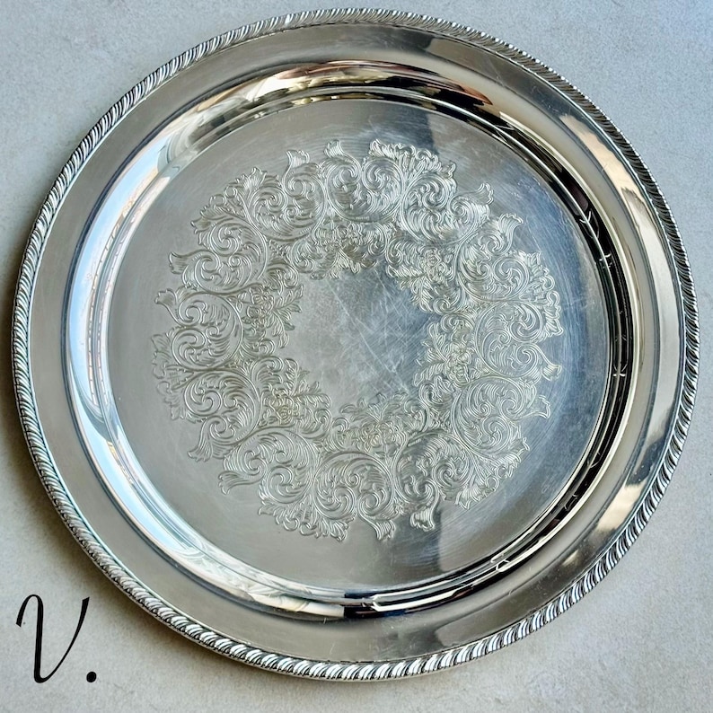 May include: A round silver-toned serving tray featuring a polished finish and a decorative engraved scrollwork pattern in the center. The rim of the tray is detailed with a textured rope-style border. The letter V is printed in the bottom left corner.
