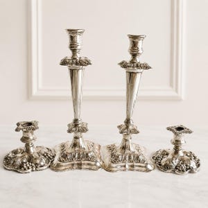 May include: A set of four silver-toned metal candle holders featuring two tall, slender candlesticks and two shorter, wide-based candle stands. Each piece is decorated with intricate embossed grape and vine leaf patterns on the square bases.