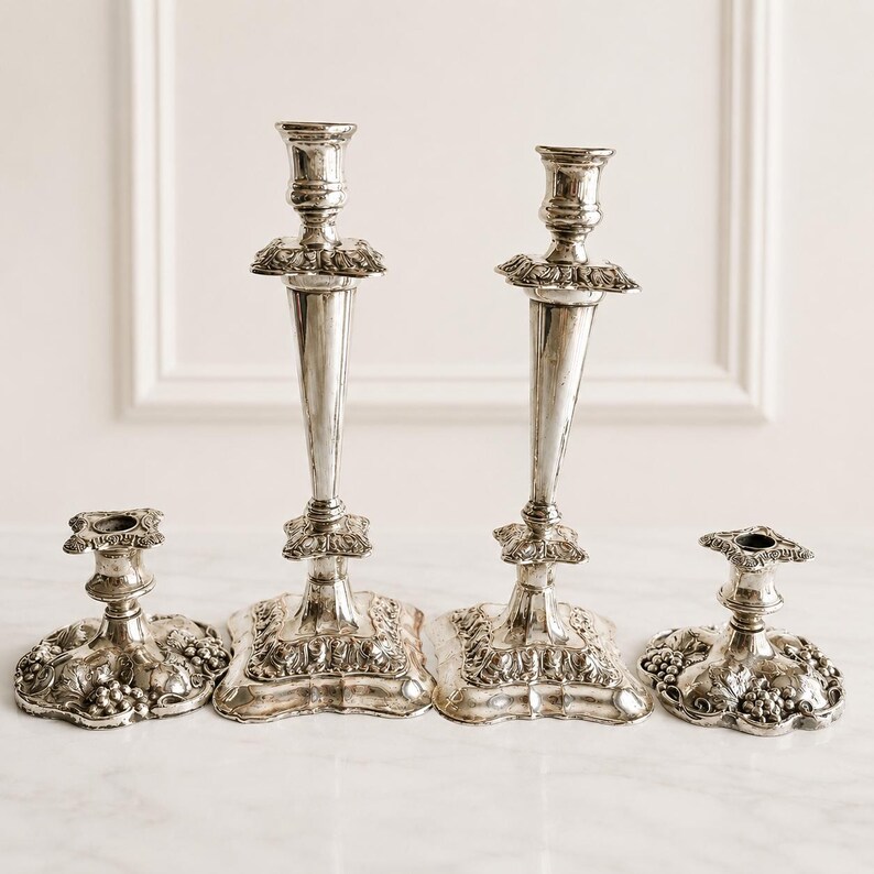 May include: A set of four silver-toned metal candle holders featuring two tall, slender candlesticks and two shorter, wide-based candle stands. Each piece is decorated with intricate embossed grape and vine leaf patterns on the square bases.