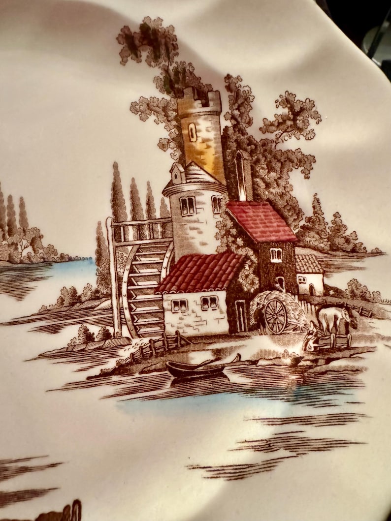 Op de afbeelding: Een wit keramieken bord met een bruine transferware-illustratie van een watermolen met een hoge toren, gebouwen met rode daken, een waterrad, een kleine boot op het water en een paard en wagen bij de rivieroever.