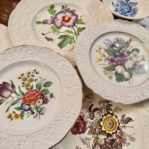 May include: A collection of ceramic plates featuring intricate embossed borders and central floral illustrations. The designs include various colorful flowers such as tulips, violets, and roses on cream-colored backgrounds with scalloped edges.