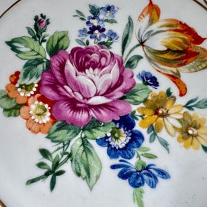 Puede incluir: Un plato de cer&aacute;mica blanco con un vibrante dise&ntilde;o floral. El arreglo presenta una gran rosa rosa de m&uacute;ltiples p&eacute;talos rodeada de flores m&aacute;s peque&ntilde;as naranjas, amarillas y azules, con hojas verdes. Borde dorado.