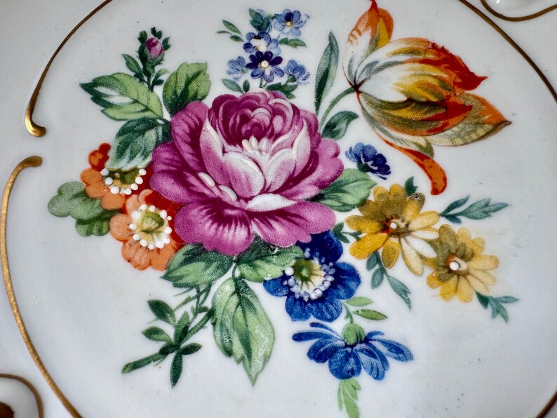 Puede incluir: Un plato de cer&aacute;mica blanco con un vibrante dise&ntilde;o floral. El arreglo presenta una gran rosa rosa de m&uacute;ltiples p&eacute;talos rodeada de flores m&aacute;s peque&ntilde;as naranjas, amarillas y azules, con hojas verdes. Borde dorado.
