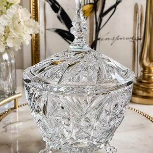 May include: A clear, cut-glass candy dish with a matching lid featuring a pointed finial. The bowl has a rounded body with intricate starburst and diamond patterns, supported by three small, curved feet. The glass is transparent and reflective.