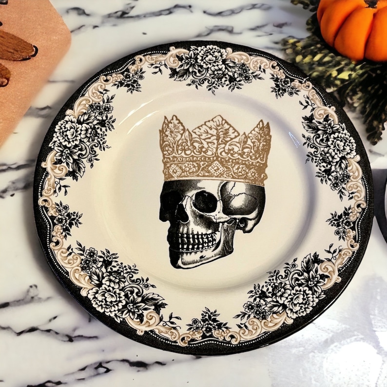 Puede incluir: Un plato redondo de cer&aacute;mica con una ilustraci&oacute;n central de un cr&aacute;neo humano que lleva una corona dorada. El borde est&aacute; decorado con un patr&oacute;n floral en blanco y negro acentuado con volutas beige sobre un fondo de color crema.
