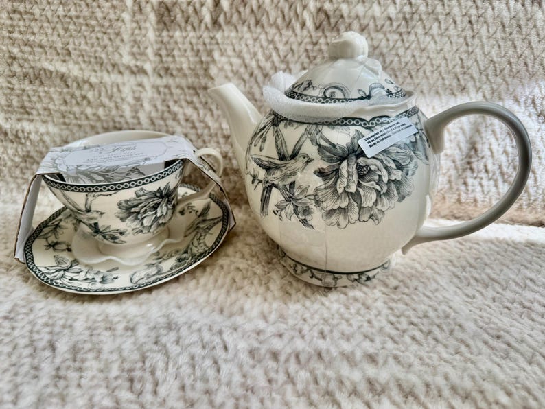 K&ouml;nnte beinhalten: Wei&szlig;es Keramik-Teeservice mit Blumen- und Vogelmuster. Das Set beinhaltet eine Teekanne mit grauem Griff und Deckel, eine Teetasse und eine Untertasse. Das Design ist in Schwarz und Wei&szlig; gehalten. Das Set steht auf einer strukturierten Oberfl&auml;che.