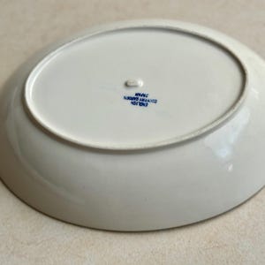 May include: An oval, white ceramic plate with a slightly raised rim. The underside features blue text that reads "ENGLISH COUNTRY GARDEN JAPAN". The plate has a small, raised foot and is likely for serving food.