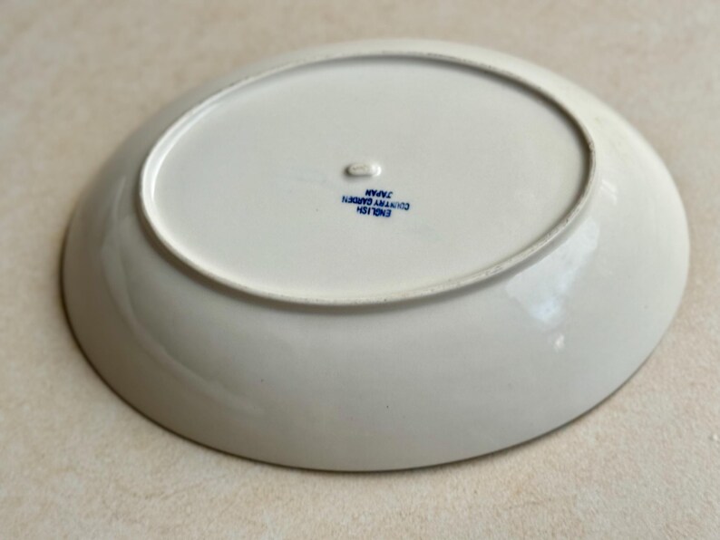 May include: An oval, white ceramic plate with a slightly raised rim. The underside features blue text that reads "ENGLISH COUNTRY GARDEN JAPAN". The plate has a small, raised foot and is likely for serving food.