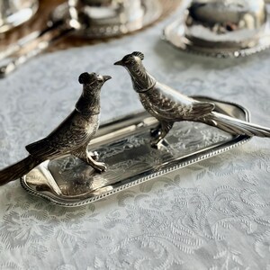 May include: Two silver pheasant figurines, perched on a rectangular silver tray with ornate detailing. The birds face each other, showcasing intricate feather and beak details. The tray sits on a white patterned tablecloth.