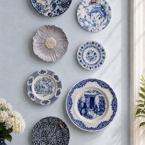 Puede incluir: Una colecci&oacute;n de siete platos decorativos de cer&aacute;mica expuestos en una pared. Los platos presentan diversos motivos florales, paisaj&iacute;sticos y de aves en azul y blanco, junto a un plato gris claro liso con un dise&ntilde;o texturizado de p&eacute;talos.