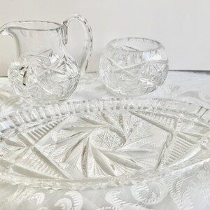May include: A clear glass set consisting of a small creamer pitcher with a handle, a matching open sugar bowl, and an oval serving tray. Each piece features intricate pressed geometric starburst and fan patterns throughout the glass surface.